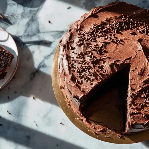 A partially served chocolate birthday cake with sprinkles on a gold board, next to a slice on a plate with a spoon, all on a marble surface.