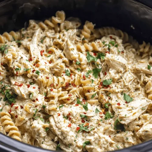 Creamy Garlic Parmesan Crockpot Chicken Pasta 2 Close-up of creamy Garlic Parmesan Crockpot Chicken with fusilli pasta, garnished with red chili flakes and fresh green herbs in a dark slow cooker.