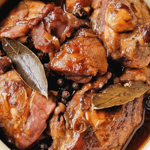Close-up of a rich Slow Cooker Chicken Adobo in a light ceramic bowl, garnished with bay leaves and whole black peppercorns.