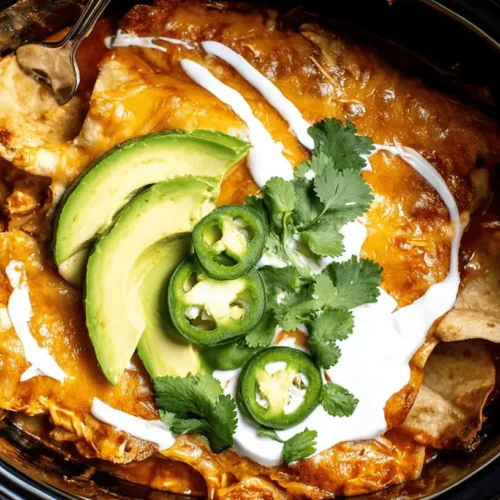 Overhead shot of hearty slow cooker chicken enchiladas, bubbling with melted cheese and topped with fresh avocado, jalapeños, sour cream, and cilantro.