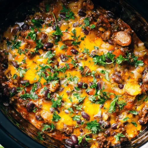 Top-down view of a hearty Slow Cooker Cowboy Casserole with beans, sausage, melted cheddar cheese, and fresh herbs simmering in a black slow cooker on a kitchen counter.