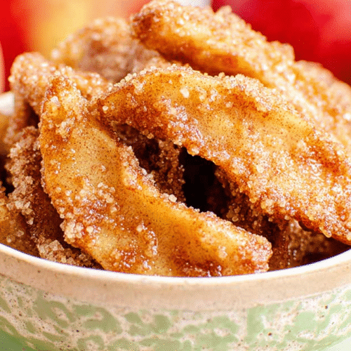 simple Air Fryer Apple Fries taken with an old camera, golden-brown apple slices, glistening brown sugar cinnamon glaze, coarse sugar crystals, textured cream ceramic bowl, light green decorative rim plate, soft-focus red apples background, soft warm light, shallow depth of field.