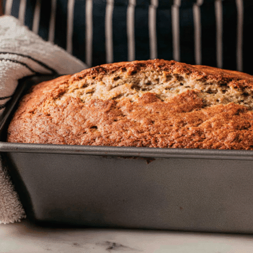 simple sourdough banana bread taken with an old camera, freshly baked loaf, rich golden-brown crust, cracked texture, warm, dark metal loaf pan, held by person, striped apron, white terry cloth towel, light stone countertop, softly blurred kitchen background, soft lighting, homemade comfort.